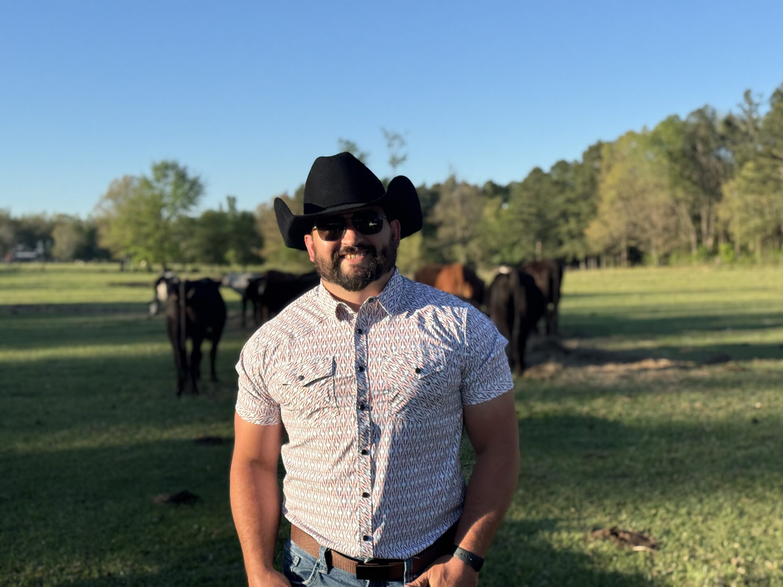 Man wearing a cowboy hat and sunglasses smiles in a green pasture with cows grazing in the background under a clear blue sky.